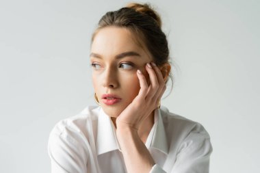 portrait of pretty young woman with natural makeup holding hand near face and looking away isolated on grey clipart