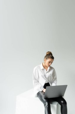 young woman in oversize shirt and black tight pants working on laptop while sitting on cube on grey background clipart