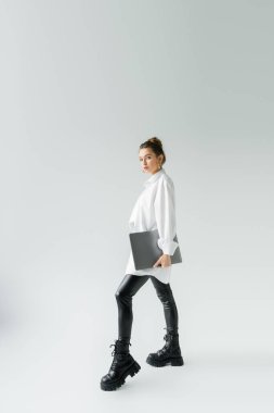 full length of stylish woman in black latex pants and rough boots posing with laptop on grey background clipart
