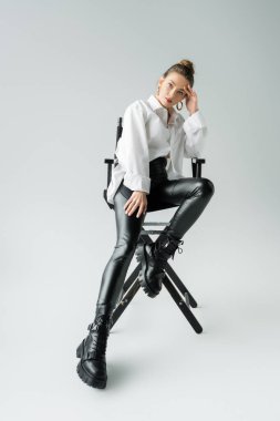 full length of stylish woman in white shirt and black pants with leather boots sitting on chair on grey background clipart