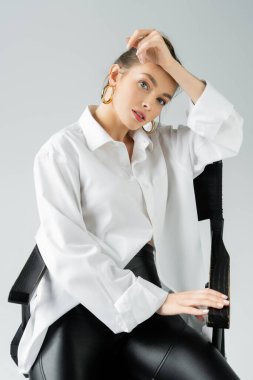 stylish woman in hoop earrings and white oversize shirt posing on chair and looking at camera isolated on grey clipart