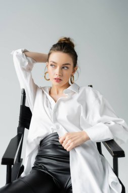 young model in white oversize shirt and black latex pants sitting on chair with hand behind head isolated on grey clipart