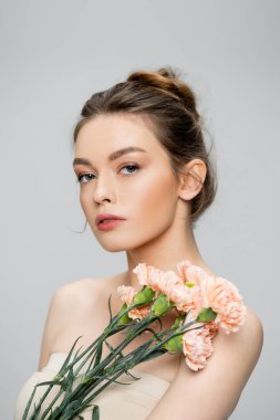 pretty young woman with bare shoulders looking at camera near bouquet of carnations isolated on grey