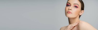 sensual woman with makeup looking at camera and touching naked shoulder isolated on grey, banner