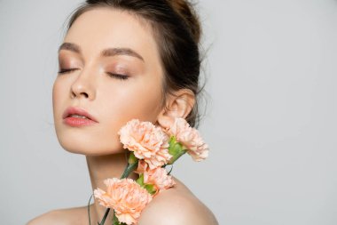 portrait of sensual young woman with closed eyes and natural makeup near peach carnations isolated on grey