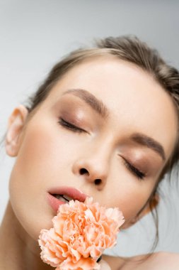 close up portrait of young woman with makeup and closed eyes posing with carnation isolated on grey
