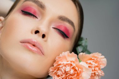 close up portrait of young woman with pink makeup posing with closed eyes near carnations isolated on grey