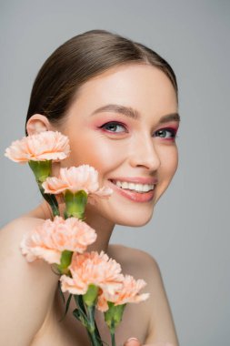 happy young woman with makeup and perfect skin smiling at camera near peach carnations isolated on grey clipart