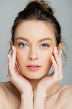 Young fair haired model with naked shoulders touching cheecks isolated on grey 