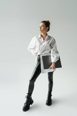 Full length of fashionable freelancer in shirt holding laptop and posing on grey 