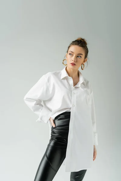 trendy woman in white shirt and black pants posing with hand on hip isolated on grey