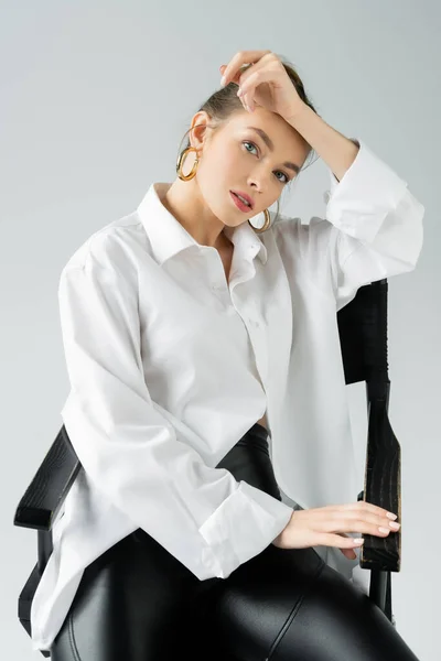 stylish woman in hoop earrings and white oversize shirt posing on chair and looking at camera isolated on grey