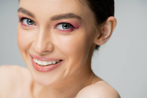 close up portrait of cheerful woman with makeup looking at camera isolated on grey