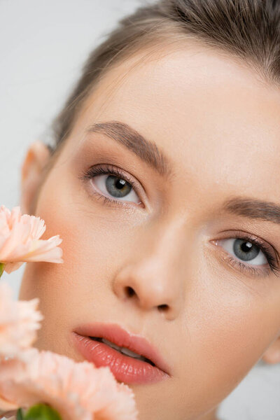 close up portrait of young woman with natural makeup looking at camera near blurred flowers isolated on grey