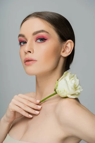 stock image sensual woman with pink makeup holding fresh rose near naked shoulder and looking at camera isolated on grey