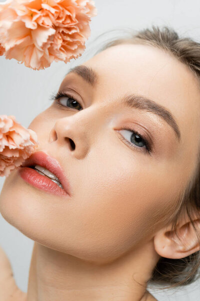 close up of charming woman with perfect face and natural makeup looking at camera near flowers isolated on grey