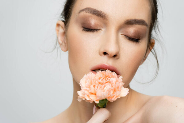 young woman with makeup and closed eyes holding peach carnation near face isolated on grey