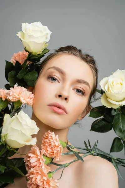young woman with natural makeup looking at camera near fresh flowers isolated on grey