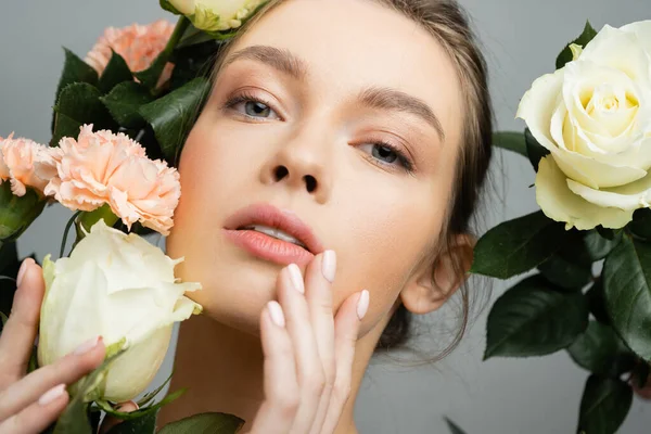 stock image portrait of sensual woman touching face while looking at camera near roses and carnations isolated on grey