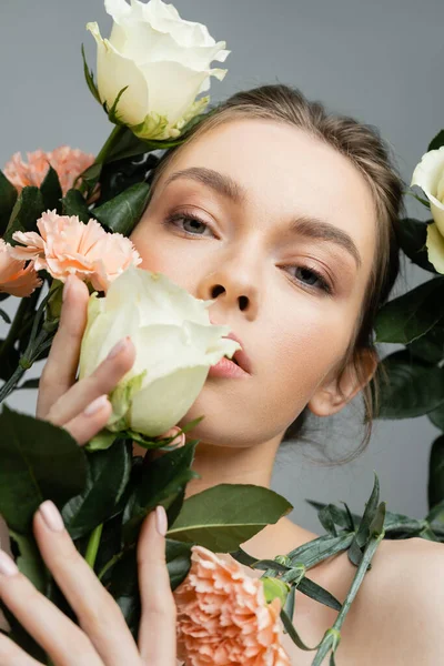 sensual woman with perfect face looking at camera while touching fresh flowers isolated on grey