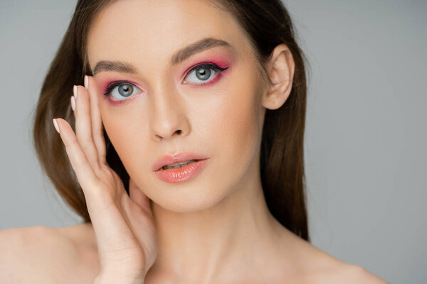 Portrait of young woman with pink makeup touching face isolated on grey 