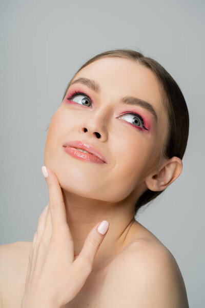 Portrait of joyful woman with pink eye shadow touching face isolated on grey 