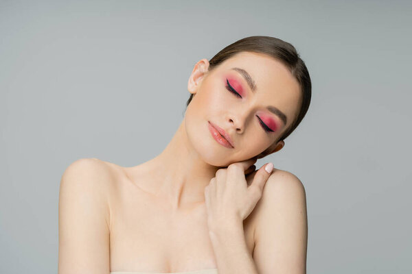 Pretty young model with pink makeup touching shoulder isolated on grey 