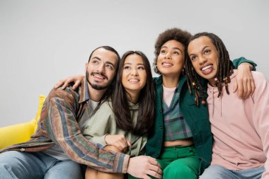 happy and trendy multicultural friends sitting and looking away isolated on grey