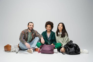 full length of stylish multicultural friends sitting with crossed legs near backpacks and smiling at camera on grey background