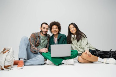 full length of multiethnic students smiling at camera while sitting with laptop on grey background clipart