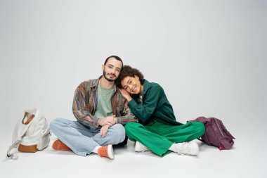 pleased interracial students sitting with crossed legs near backpacks and looking at camera on grey background