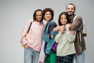 cheerful and trendy multicultural friends with backpacks posing isolated on grey