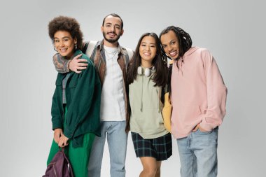smiling multicultural students in trendy hoodies and shirts looking at camera while standing with backpacks isolated on grey