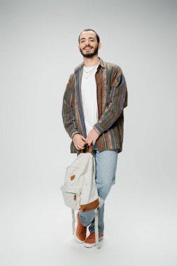 full length of cheerful bearded student in trendy outfit standing with backpack on grey background