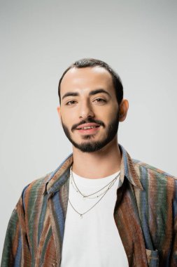 portrait of positive bearded man in trendy shirt and necklaces smiling at camera isolated on grey