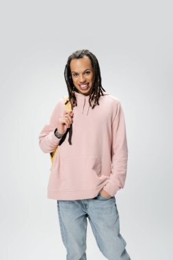 smiling multiracial student in pink hoodie holding backpack while posing with hand in pocket of blue jeans isolated on grey