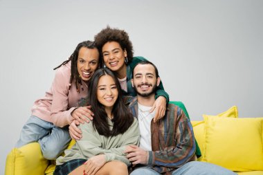 pleased multiethnic friends wearing trendy clothes and posing near yellow couch isolated on grey