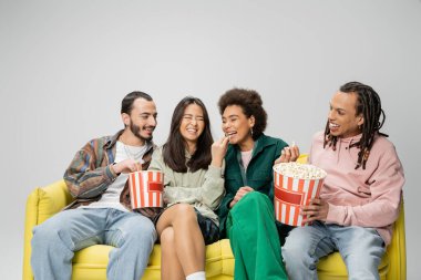 cheerful multiethnic friends in trendy clothes sitting with buckets of popcorn on yellow sofa isolated on grey