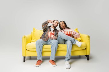full length of bearded man pointing with finger while watching movie with multiracial friend with popcorn on grey background clipart