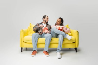 full length of stylish multiethnic men smiling at each other while sitting with buckets of popcorn on yellow couch on grey background