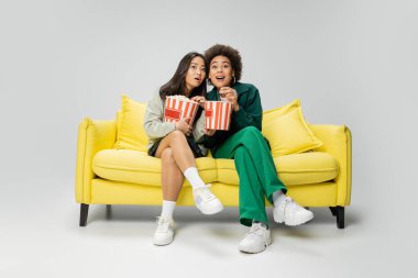 full length of astonished interracial women holding popcorn while watching movie on yellow couch on grey background