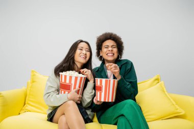 excited multiethnic women sitting on yellow couch with buckets of popcorn and watching movie isolated on grey