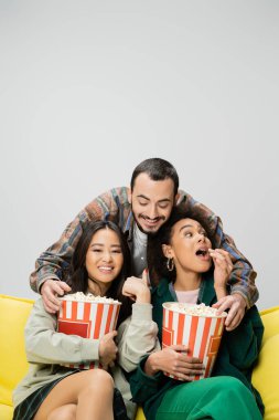 cheerful bearded man having fun while reaching popcorn near interracial women watching movie isolated on grey