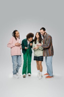 full length of bearded man showing mobile phone to laughing multiethnic friends on grey background