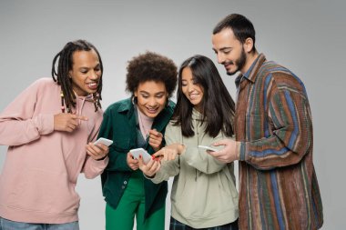 cheerful asian woman pointing at smartphone near trendy multiethnic friends isolated on grey clipart