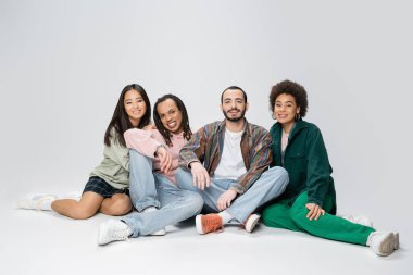 full length of positive multiethnic friends in trendy casual clothes sitting on grey background