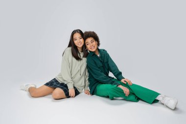 full length of happy multiethnic women in stylish casual clothes sitting on grey background