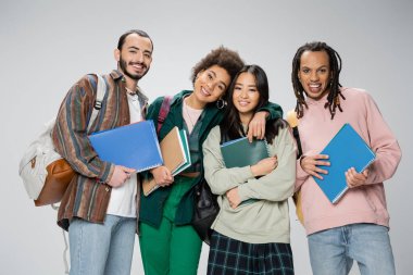 happy multiethnic students with notebooks and backpacks smiling at camera isolated on grey