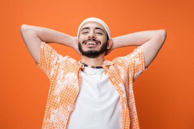 cheerful bearded man in fashionable attire posing with hands behind head isolated on orange