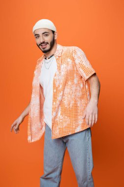 young bearded man in trendy casual attire smiling at camera while standing isolated on orange clipart